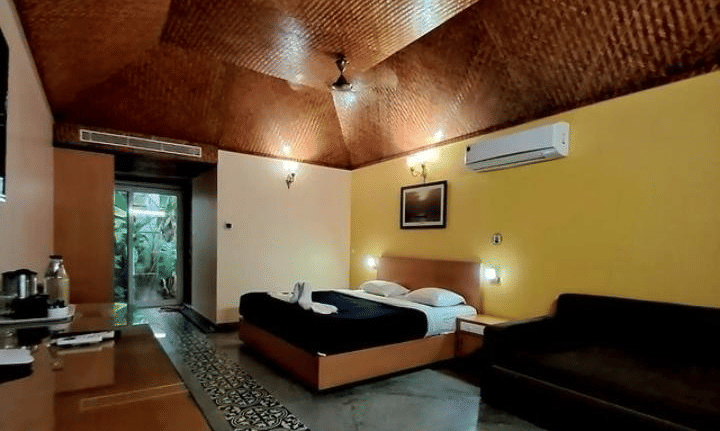 A cosy room with a queen bed, a sofa, wooden furnished ceiling and table with marble floor at Paradise Lagoon Resort, Udupi.