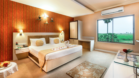 A brightly lit hotel room with a double bed with towel swans, a wood-panelled wall, and a large window with a view at Regency Tuticorin by GRT Hotels.