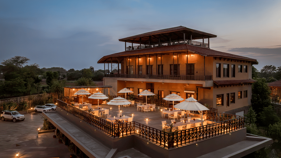 The multi-story resort building at AamaGhati Wildlife Resort with an illuminated outdoor dining deck featuring white patio umbrellas.