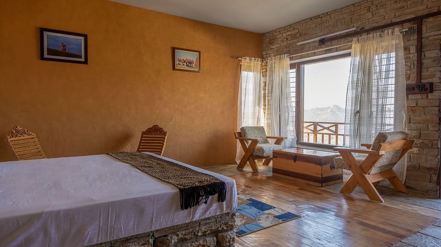 Rustic room with a bed, wooden chairs, stone wall, and a window with a mountain view. - Blusalzz Homes The Himalayan Bungalow, Almora