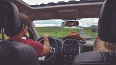 An inside view of a car with a driver and passenger in the front on a road trip with greenery in view.