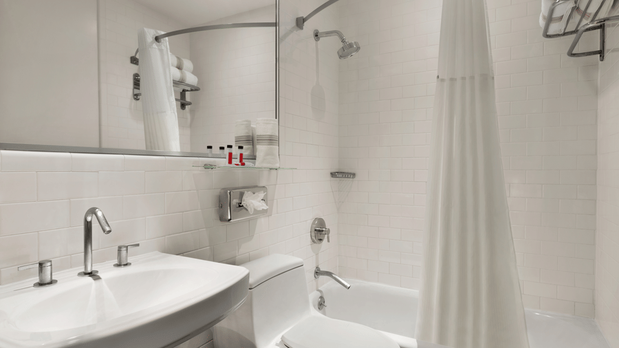 Bathroom at Night Hotel Broadway featuring a walk in shower sink toilet wall mounted fixtures white tiles and a clean minimal interior.