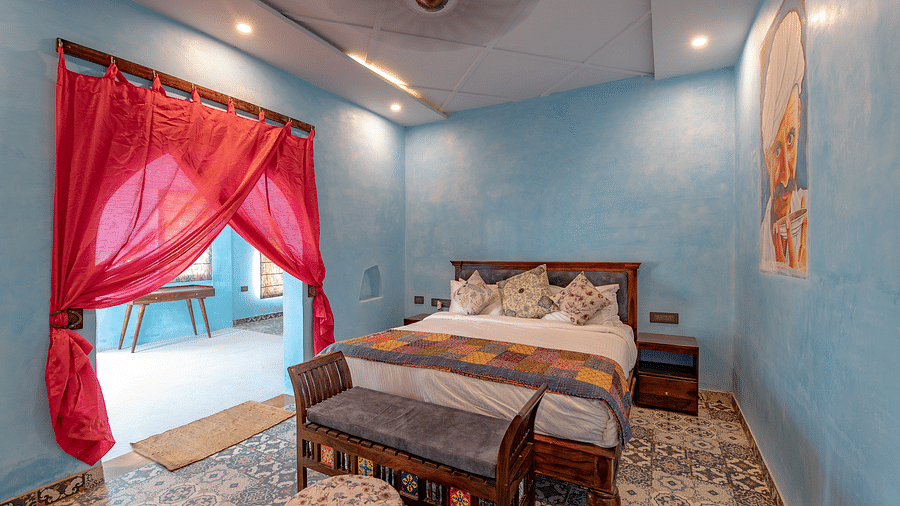 An image of a bedroom with a king size bed, a bedside table, a fan running on a ceiling, a tool near the, and an open entrance giving a sneak peak of the outdoor - Karwaan Jaisalmer