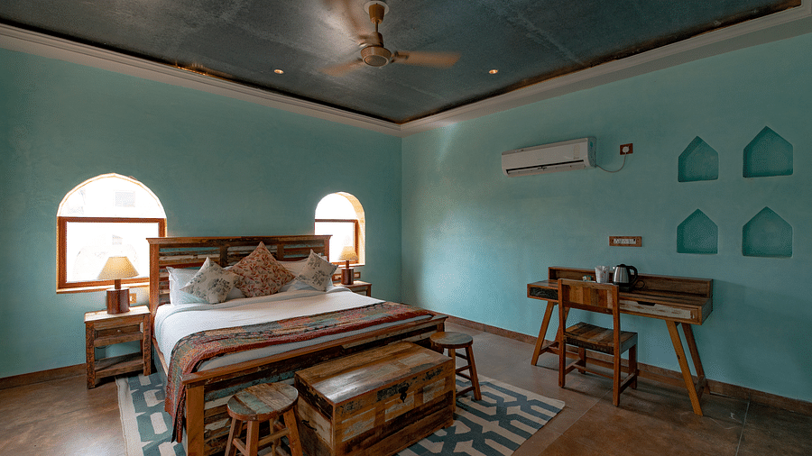 An image of a bedroom with double size bed, bedside table on either side, two open windows, and a table on one corner of the room to place various items - Karwaan Jaisalmer
