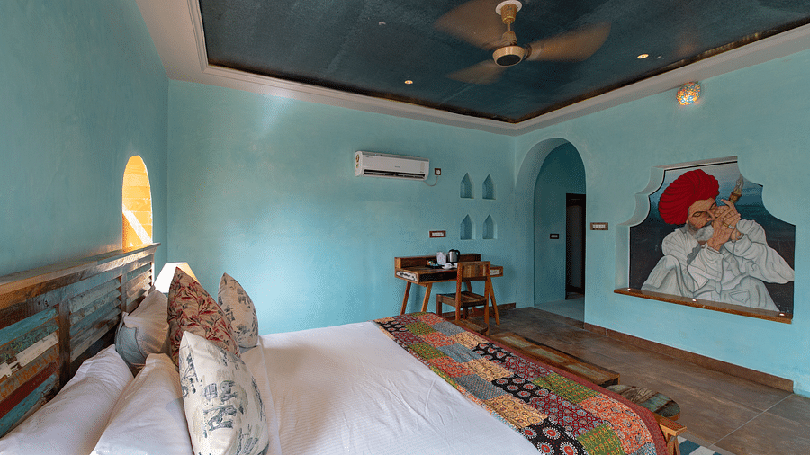 An image of a bedroom with double size bed, bedside table on either side, two open windows, and a table on one corner of the room to place various items - Karwaan Jaisalmer