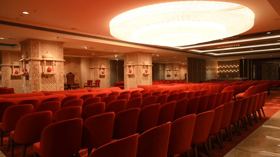 Rows of neatly arranged seats face towards the front under a large glowing ceiling light in the Lambic banquet hall at Praveg's Grand Eulogia, Ahmedabad.