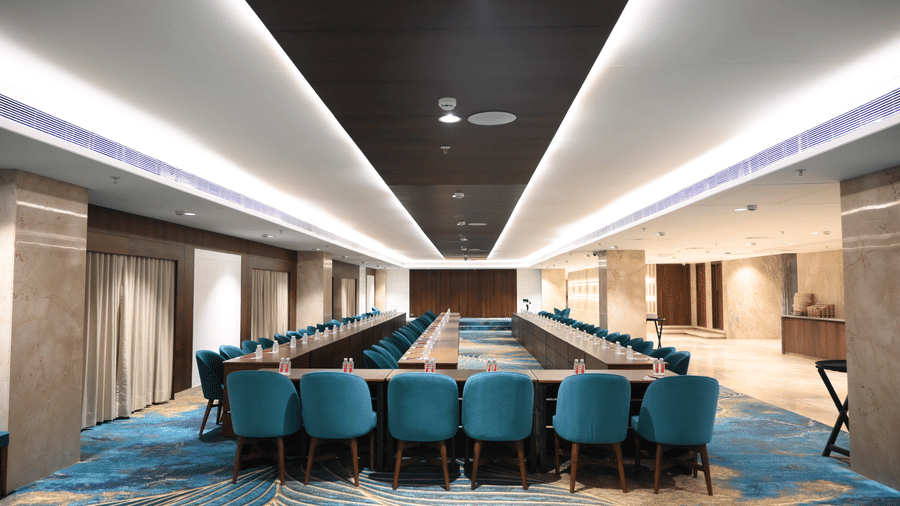 A large conference table with upholstered chairs is set up on a patterned carpet inside the Sky Lounge at Praveg's Grand Eulogia, Ahmedabad.