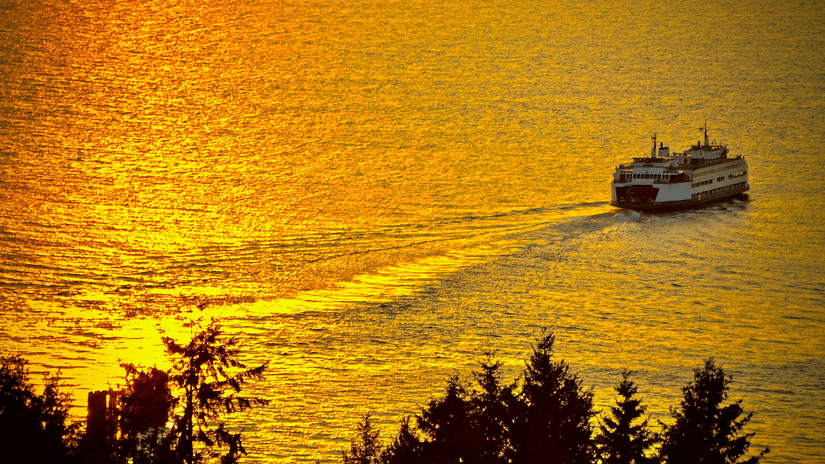 A view from the land of a ferry travelling on a waterbody during sunset.