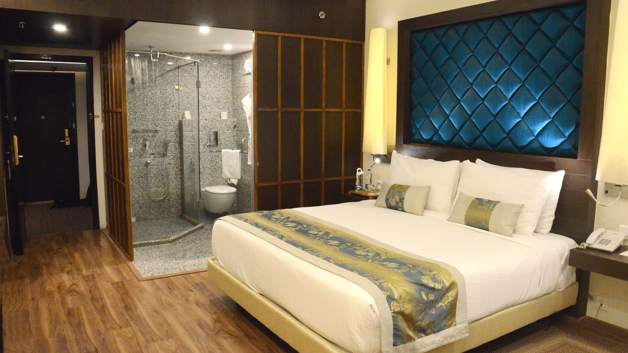 A room with a neatly made bed and bedside tables, warm lighting, and a doorway leading to a modern washroom at Clarks Avadh, Lucknow | Rooms with Gomti River View.