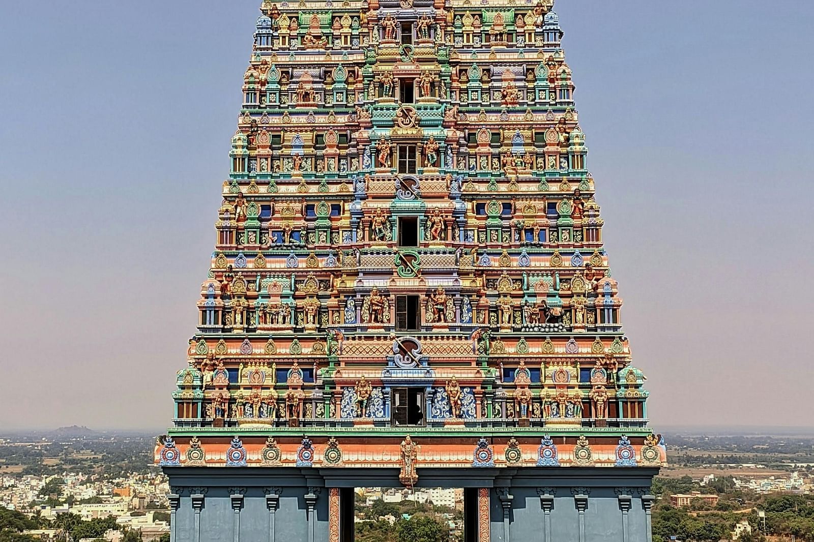 A tall, colourful South Indian temple richly adorned with vibrant sculptures of deities, mythological figures, and ornate carvings, rising high above the distant cityscape.