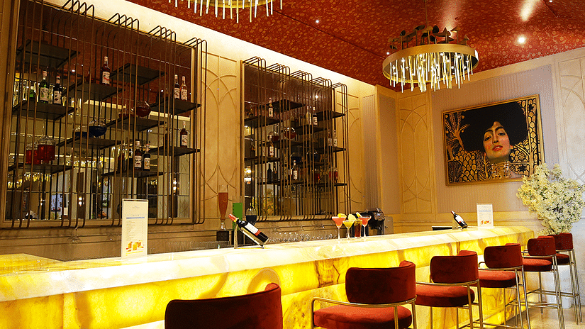 A long shot of a bar with a brightly lit countertop and red bar stools. Mirrored panels and decorative lighting enhance the ambiance. Shelves filled with bottles extend along the back wall = The Gaurs Sarovar Premiere