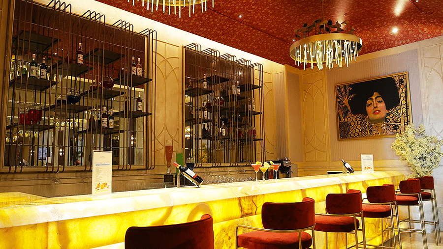 A long shot of a bar with a brightly lit countertop and red bar stools. Mirrored panels and decorative lighting enhance the ambiance. Shelves filled with bottles extend along the back wall = The Gaurs Sarovar Premiere