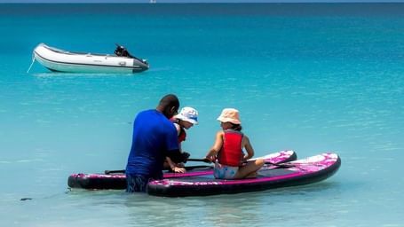A man and two kids on paddleboards in shallow turquoise water, with an inflatable boat and sailboat in the background.