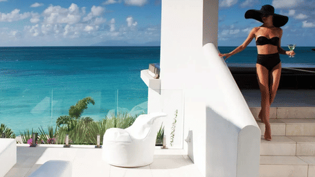 A woman in a black bikini and sun hat descends white stairs at a beachfront villa, holding a drink, with the blue sea beyond.