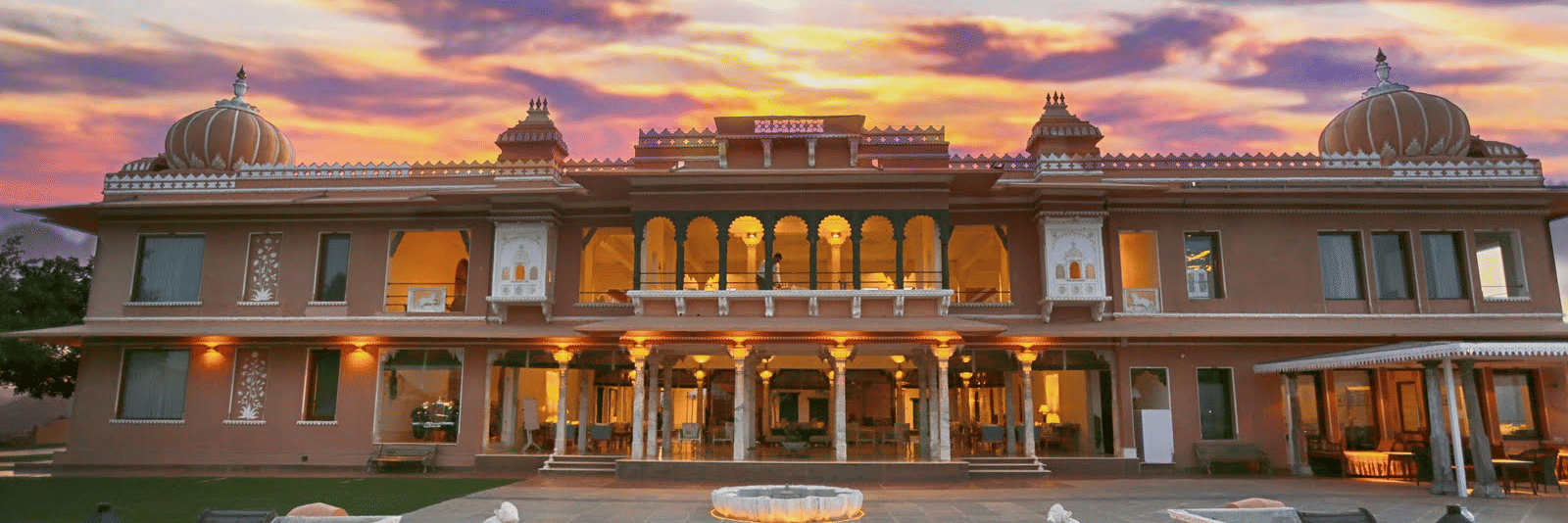 Well lit facade of Fateh Garh, Udaipur featuring a double storey building with palace style architecture. There is a flight of stairs leading to a front lawn with fountain and then the entrance of the hotel.