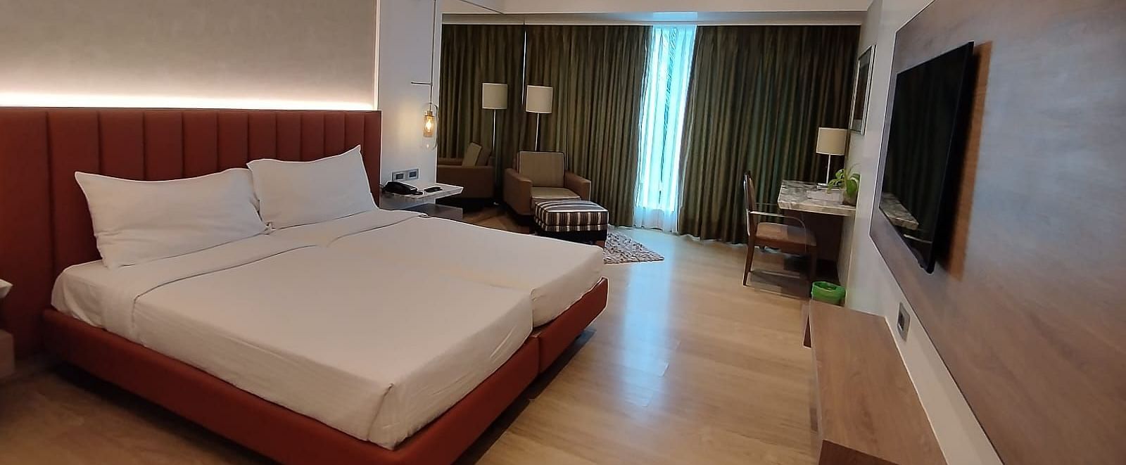 A well-lit executive bedroom in Mirage Hotel, featuring twin beds, decorative head-rest, lounge chairs, a wooden panelled entertainment unit and a large wall to wall window with closed drapes.