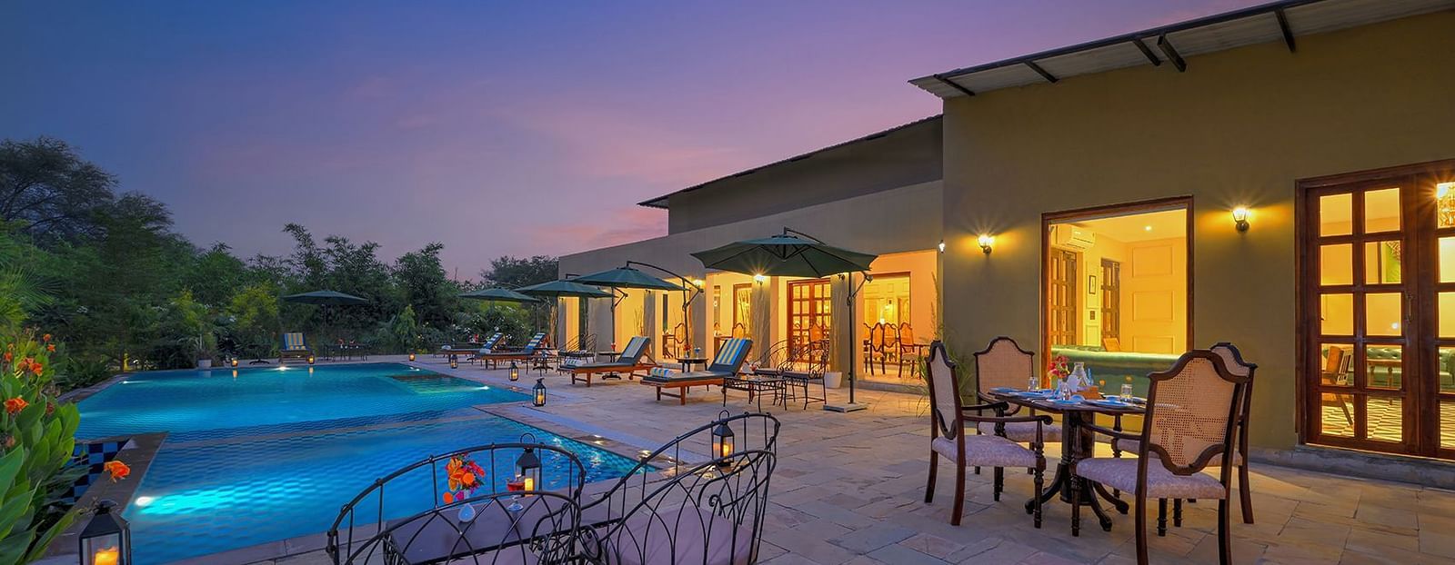 Poolside seating area with cushioned chairs and dining tables, surrounded by greenery and sun loungers under large umbrellas, illuminated by lanterns at dusk - Singhvilas, Ranthambore