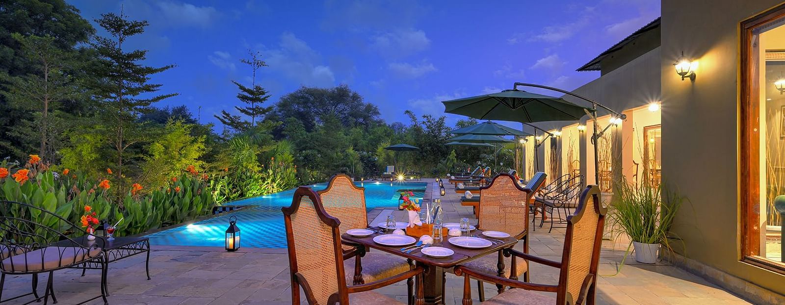 Outdoor dining setup with a wooden table and cushioned chairs near a swimming pool, surrounded by greenery, lounge chairs, and large green umbrellas under a twilight sky - Singhvilas, Ranthambore
