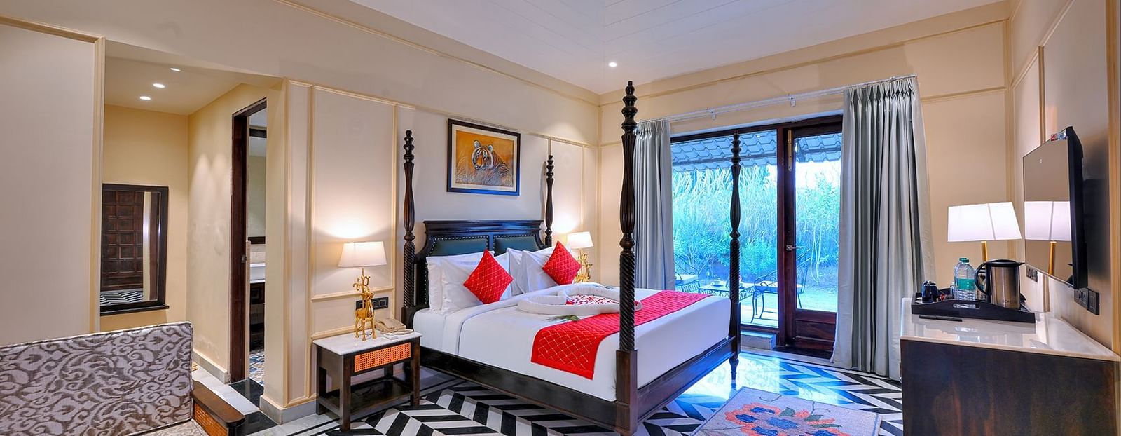 Premium Suite Cottage at Singhvilas Ranthambore features a four-poster bed with red accents, a balcony, a black and white zigzag patterned floor, comfortable seating, a TV, and tiger-themed artwork.