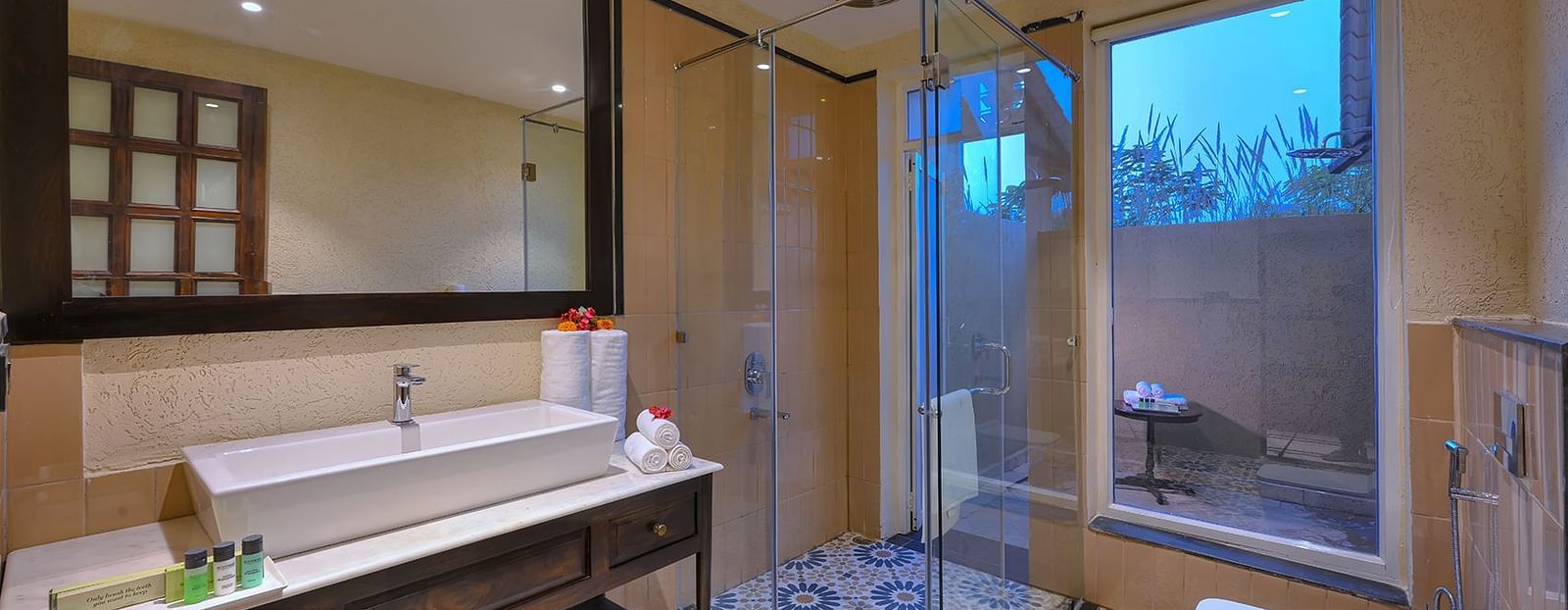 Luxurious bathroom of Superior Cottage at Singhvilas, Ranthambore, featuring a spacious vanity, glass-enclosed shower, elegant patterned flooring, and an outdoor sit-out area visible through large windows.