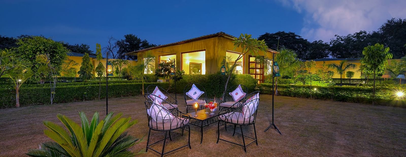 A serene garden at night featuring a table and chairs surrounded by lush greenery and soft lighting - Singhvilas, Ranthambore