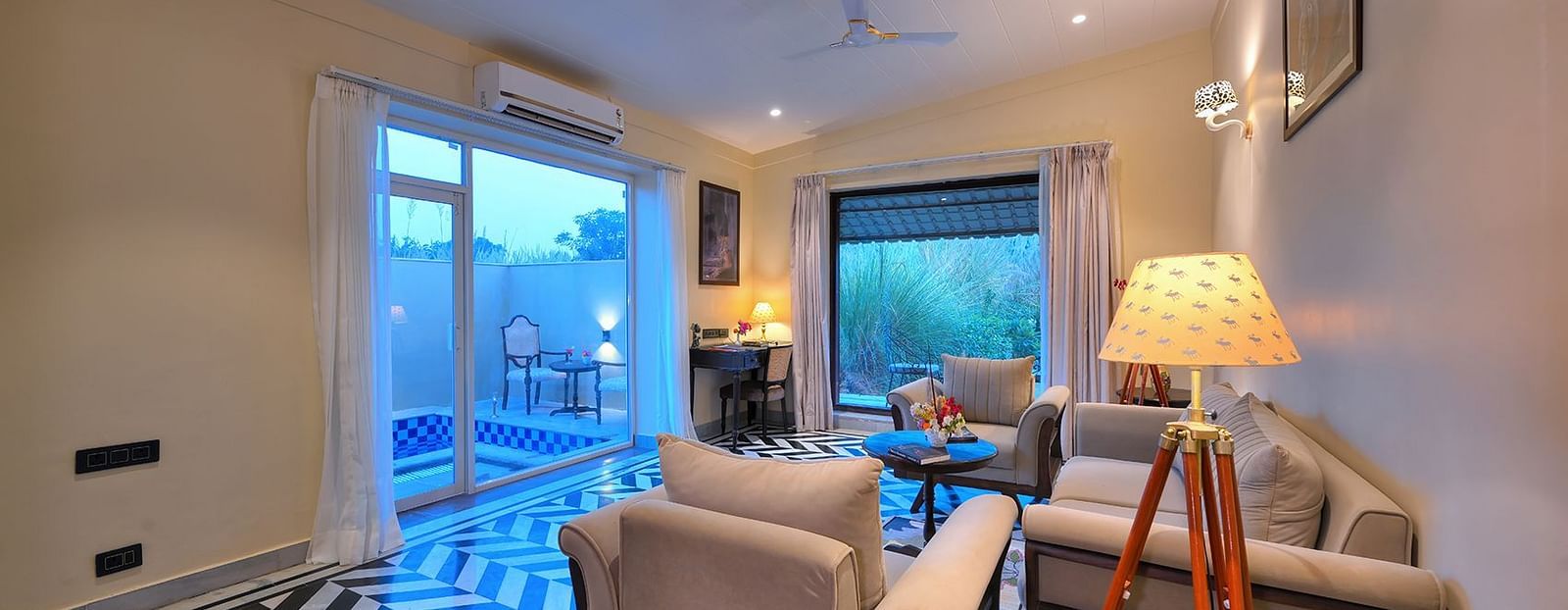 A stylish living area of Premium Suite Cottage featuring two beige armchairs, a light-coloured sofa, a tripod floor lamp with a patterned shade, and glass doors leading to a balcony with a blue-tiled pool - Singhvilas, Ranthambore