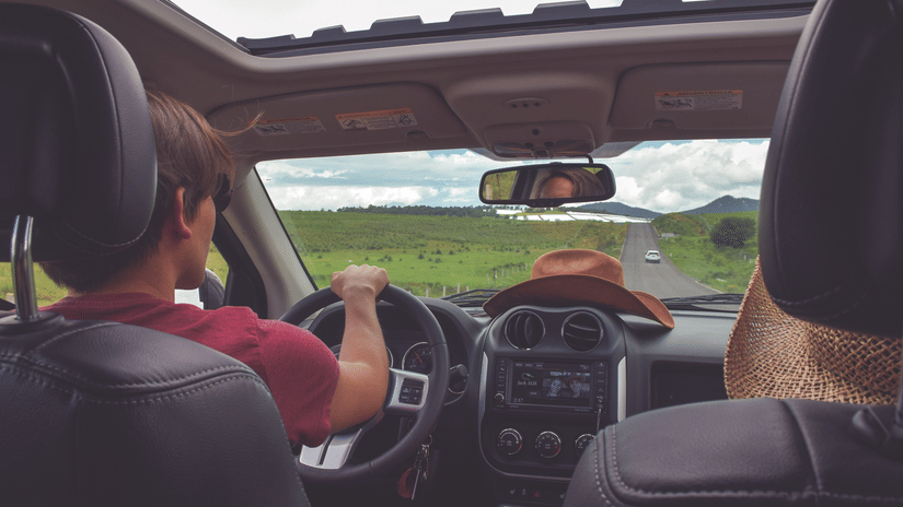 An inside view of a car with a driver and passenger in the front on a road trip with greenery in view.