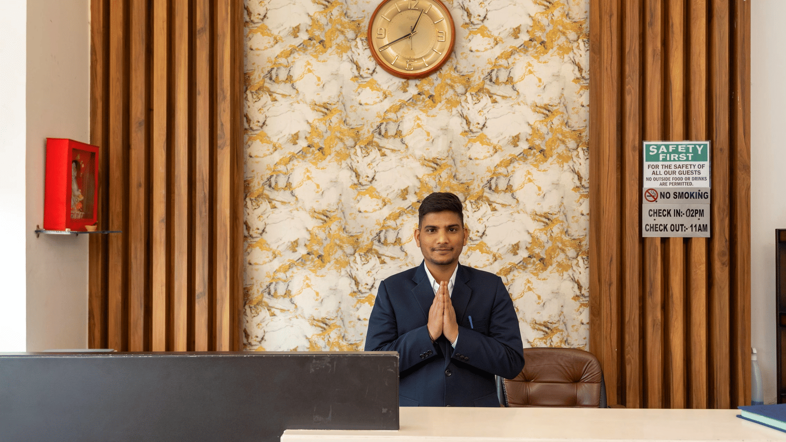 A man standing behind a reception desk with a wall clock and textured wallpaper at Perfectstayz Premium Shanti Heritage, Haridwar