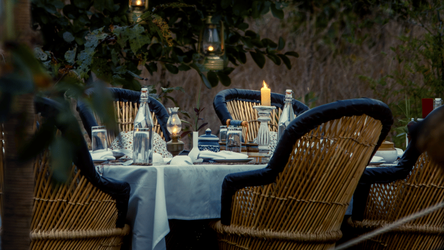 A vertical shot of a dining table surrounded by wicker chairs set up for a dimly lit dinner under trees at night - Umaria Safari Camp