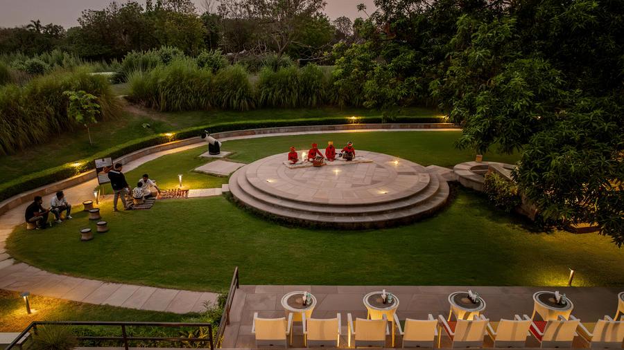 Aerial view of the lawn at AamaGhati Wildlife Resort with a circular stone performance stage, folk musicians seated on central stage, and an outdoor seating terrace.