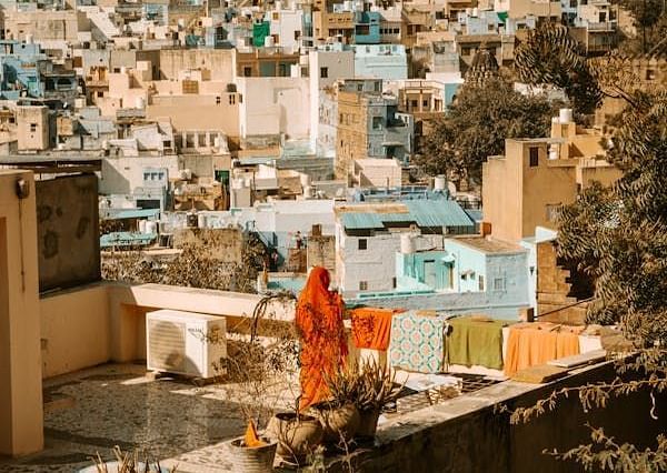 A person in bright clothing hangs laundry on a rooftop overlooking a densely populated town with a hillside in the background.