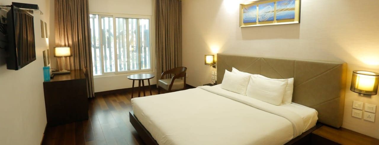 Spacious room with a large double bed, bedside lamps, wall art above the headboard, and a window with curtains pulled to the side at The Hamuse Hotel, Kodaikanal.