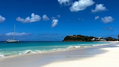 A sailboat drifting on clear turquoise water next to a wide, pristine white sand beach under a bright blue sky with fluffy clouds.