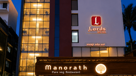 Night view of an illuminated multi-story hotel building featuring a glass-paneled stairwell, warm exterior lighting, and the ground-level entrance to Manorath Fine Dining Restaurant - Lords Eco Inn Belagavi