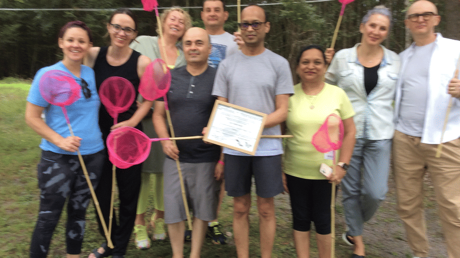 A group of nine people standing on grass outdoors, holding pink nets on sticks, with trees in the background and one person holding a certificate at YO1 Longevity & Health Resorts, Catskills