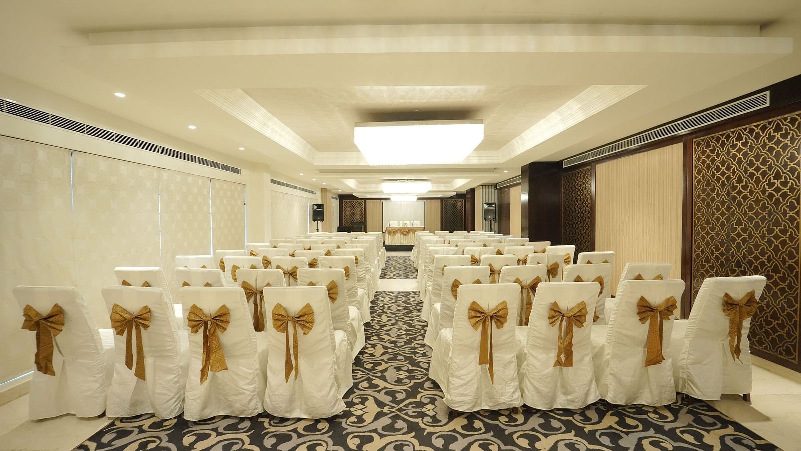 A banquet hall with rows of chairs draped in white covers and gold bows, ready for an event - Fragrant Nature Cochin