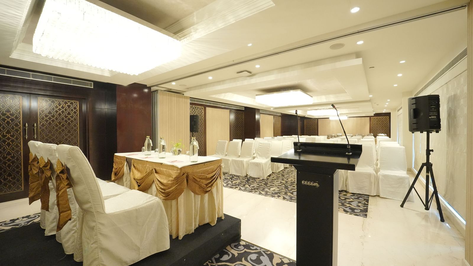 A conference room setup with a podium, speaker system, and chairs with white covers and gold sashes - Fragrant Nature Cochin