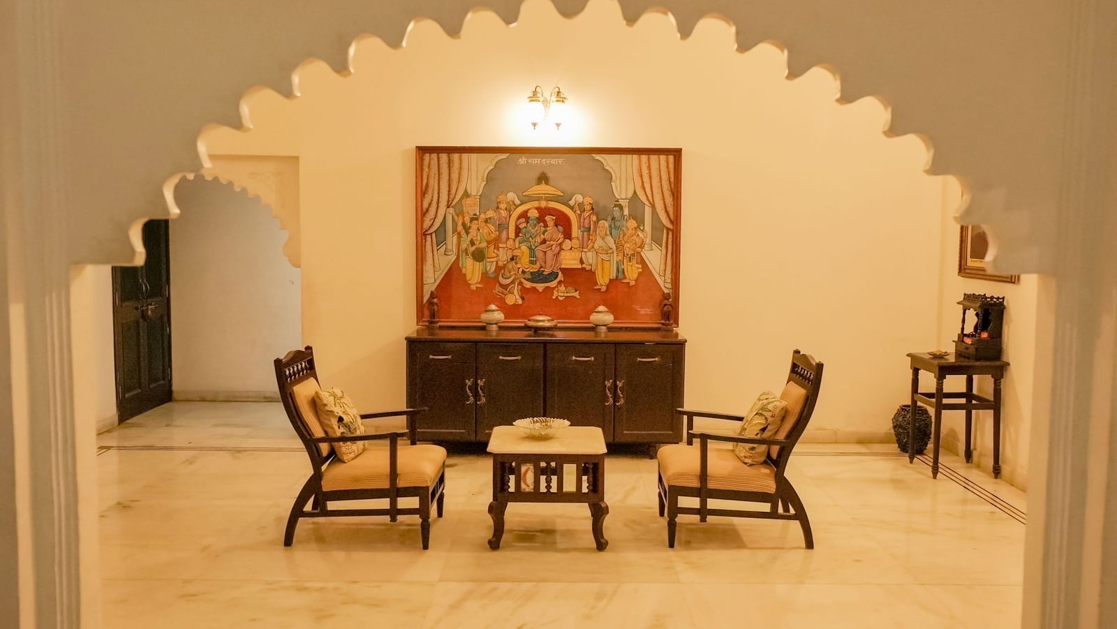 A decorative sitting area in the Terrace Suite at Mahendra Niwas. Two wooden chairs with yellow cushions face a small table. A colorful painting hangs on the wall.