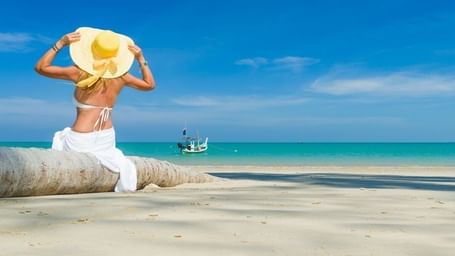 A woman sitting on a fallen trunk of a coconut tree in the sea shore, facing the expansive sea and a yacht.