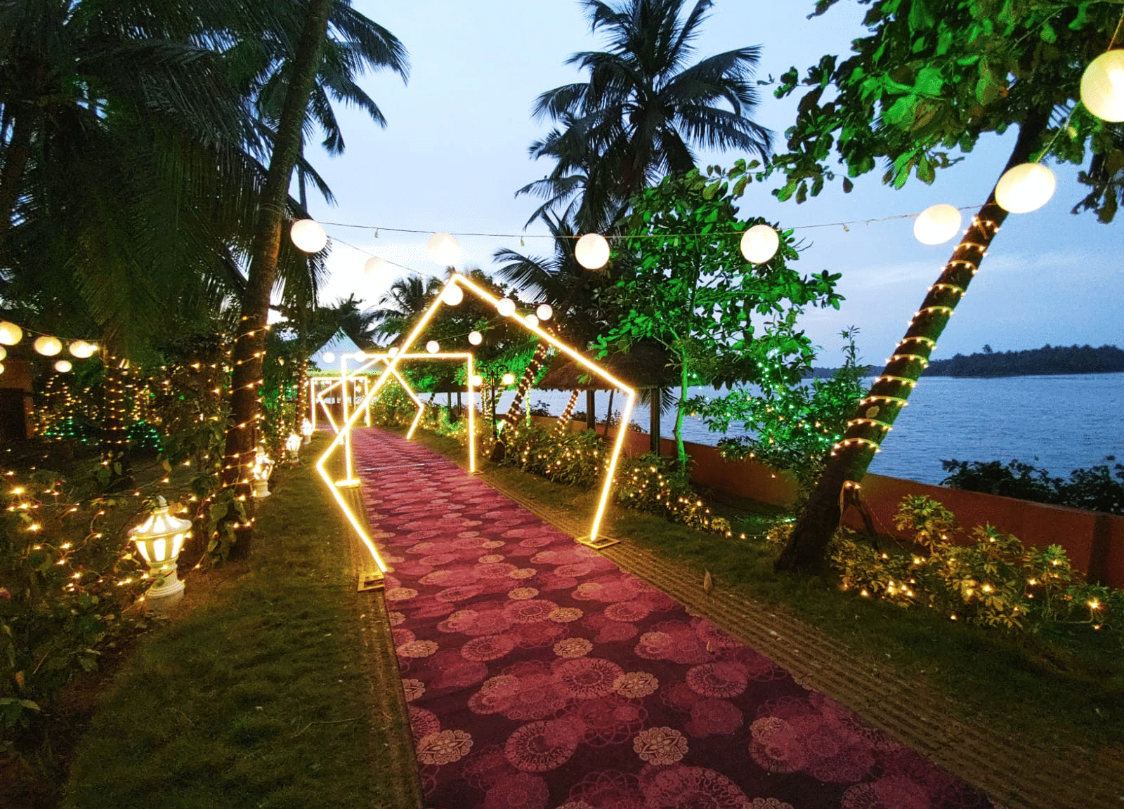 Pathway lined with lights and flowers leading towards the water with palm trees at Paradise Lagoon Resort, Udupi.