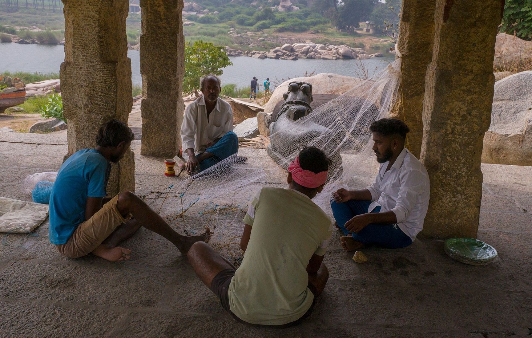 Men mending a fishing net under a stone pavilion in Hampi.