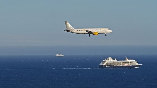 A shot that captures a ship sailing in water and plane flying in the same frame with blue sky and crystal water as the backdrop