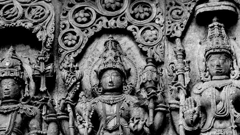 A detailed monochrome stone relief carving depicting intricate Hindu deities with elaborate headpieces and ornate floral patterns.