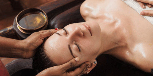 A woman lying beside a bowl of liquid while a therapist massages her head.