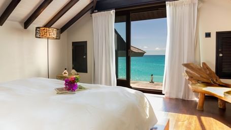 Cozy beachfront bedroom with vaulted ceilings, white linens, and open curtains revealing stunning ocean views just steps from the private terrace.