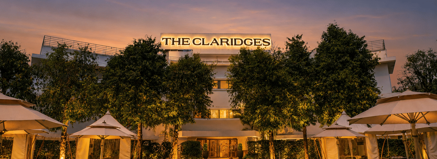 Evening view of a green hotel lawn featuring warm-lit white canopies and outdoor dining tables in front of an illuminated building at dusk - The Claridges New Delhi