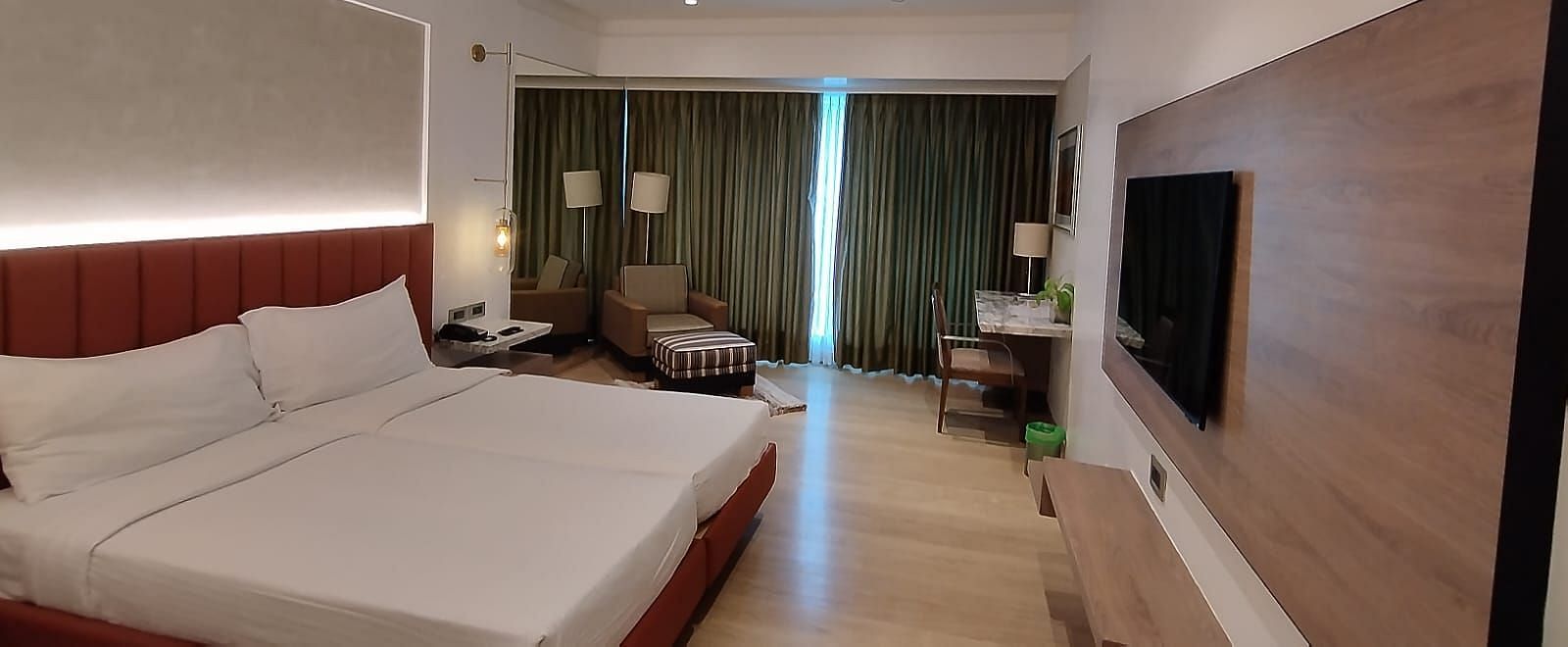 An executive bedroom in Mirage Hotel, featuring twin beds, decorative head-rest, lounge chairs, an entertainment unit, and a large wall to wall window with closed drapes.