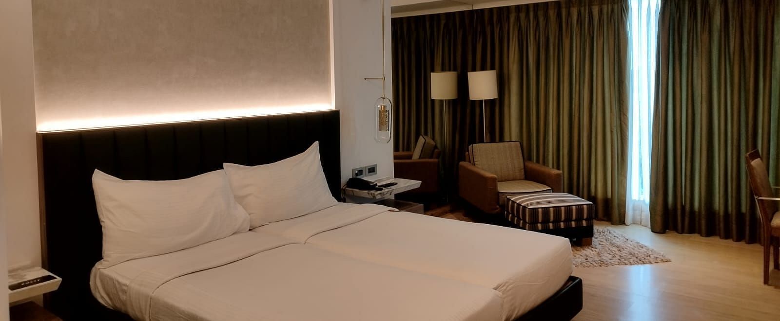 A warmly lit executive bedroom in Mirage Hotel, featuring twin beds, decorative head-rest, lounge chairs, and a large wall to wall window with closed drapes.