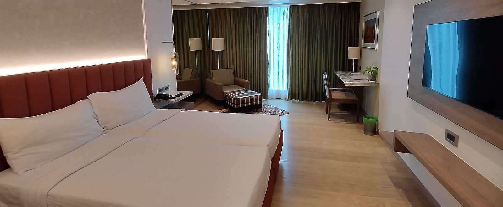 A warmly lit executive bedroom in Mirage Hotel, featuring twin beds, decorative head-rest, lounge chairs, a wooden panelled entertainment unit and a large wall to wall window with closed drapes.