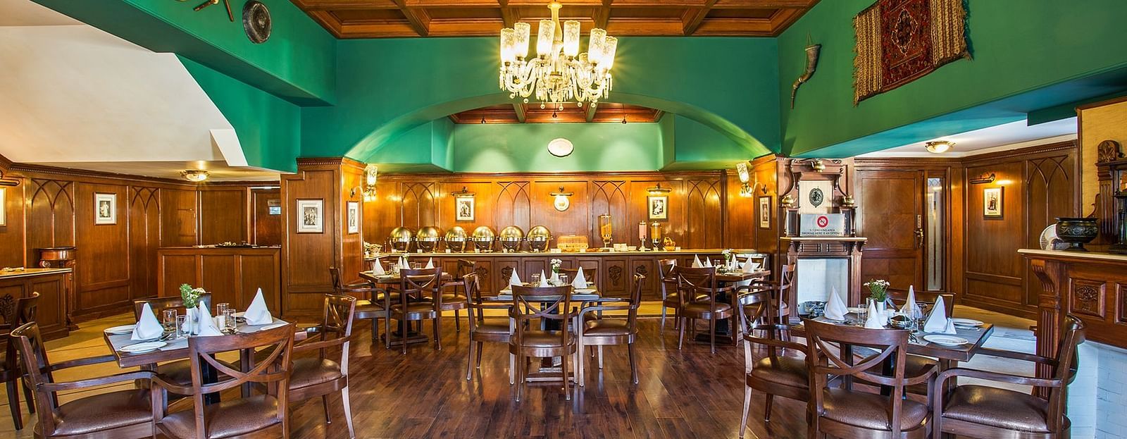 An image showing the interior of restaurant at The naini retreat, Nainital. featuring wooden decor, flooring, and a wooden bar. A large chandelier hangs from the ceiling, providing ambient lighting.